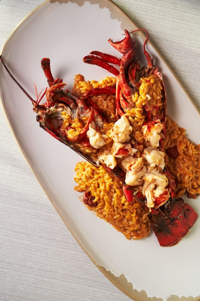 Lobster Orzo: An Elegant Seafood Dinner for Special Occasions Split lobster served with creamy orzo and chunks of lobster meat on a large white platter.