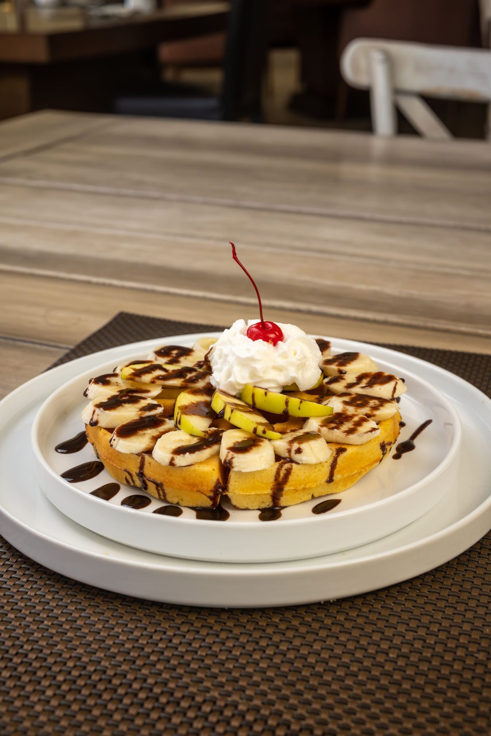 Banana chocolate waffles topped with fresh banana slices, whipped cream, chocolate drizzle, and a cherry.