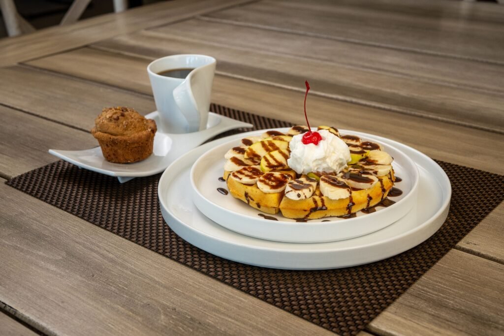 Banana chocolate waffles topped with sliced bananas, whipped cream, chocolate drizzle, and a cherry, served with coffee and a muffin.