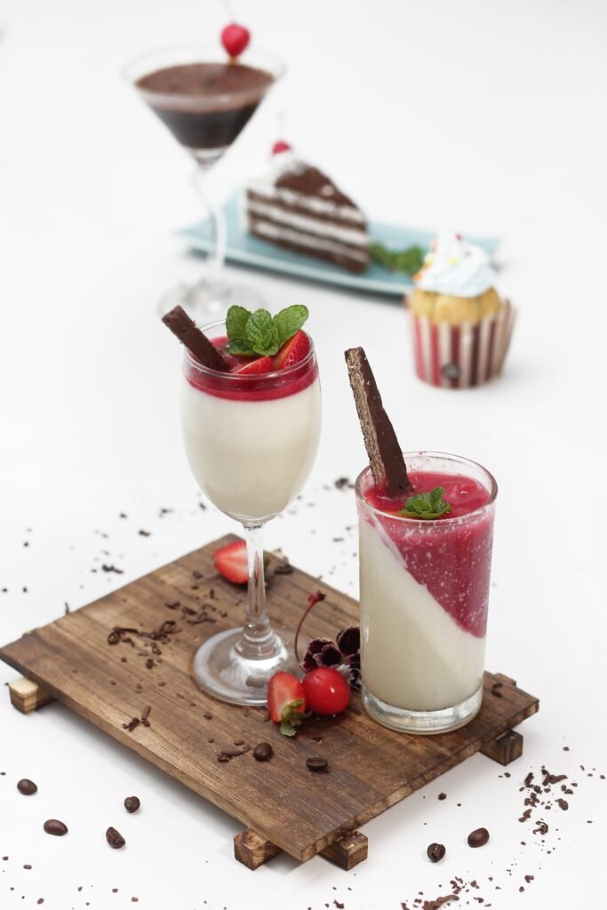 Two strawberry panna cotta desserts in glasses topped with berry sauce, mint, and chocolate wafers.