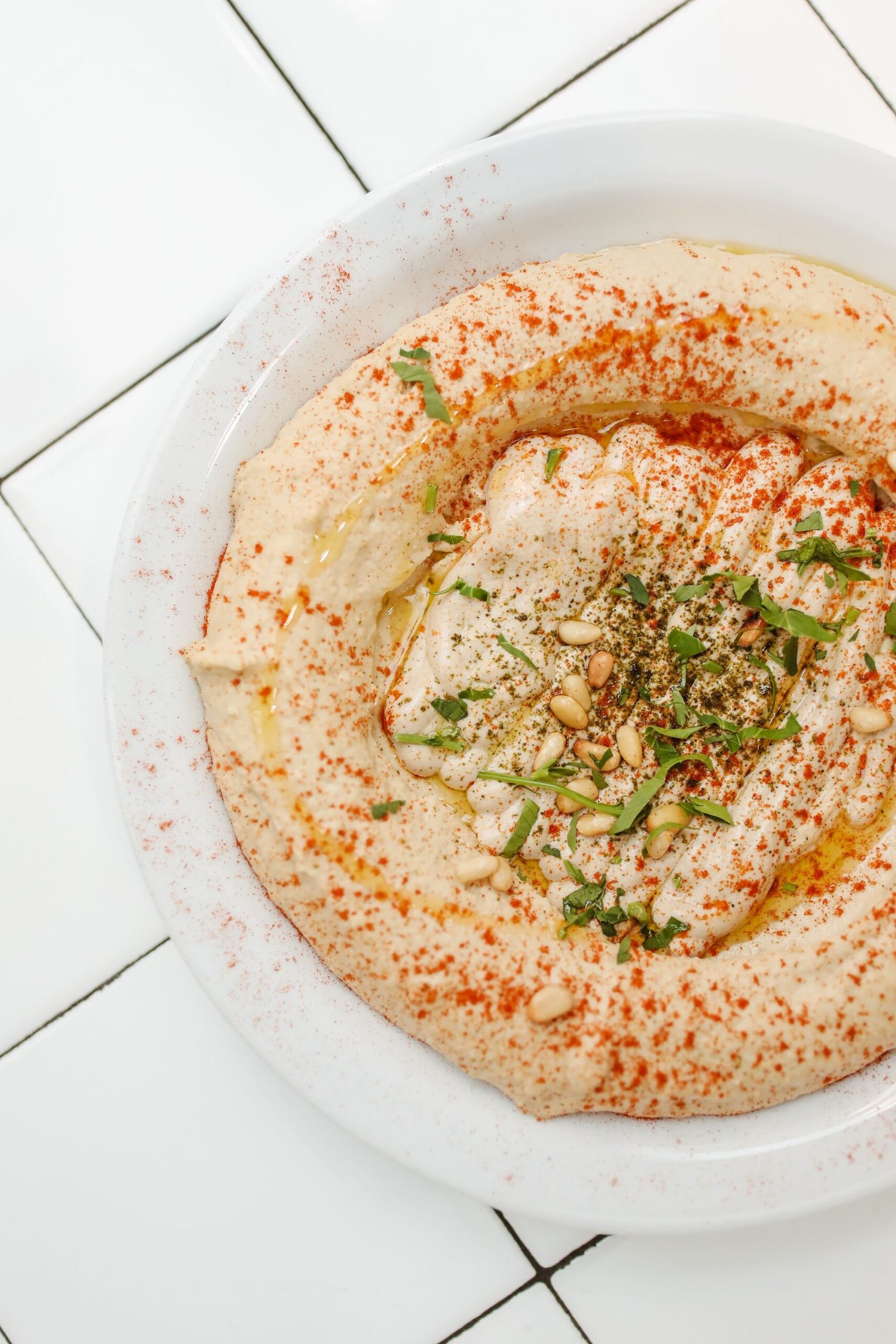 Creamy homemade hummus topped with olive oil, paprika, fresh herbs, and pine nuts in a white bowl.