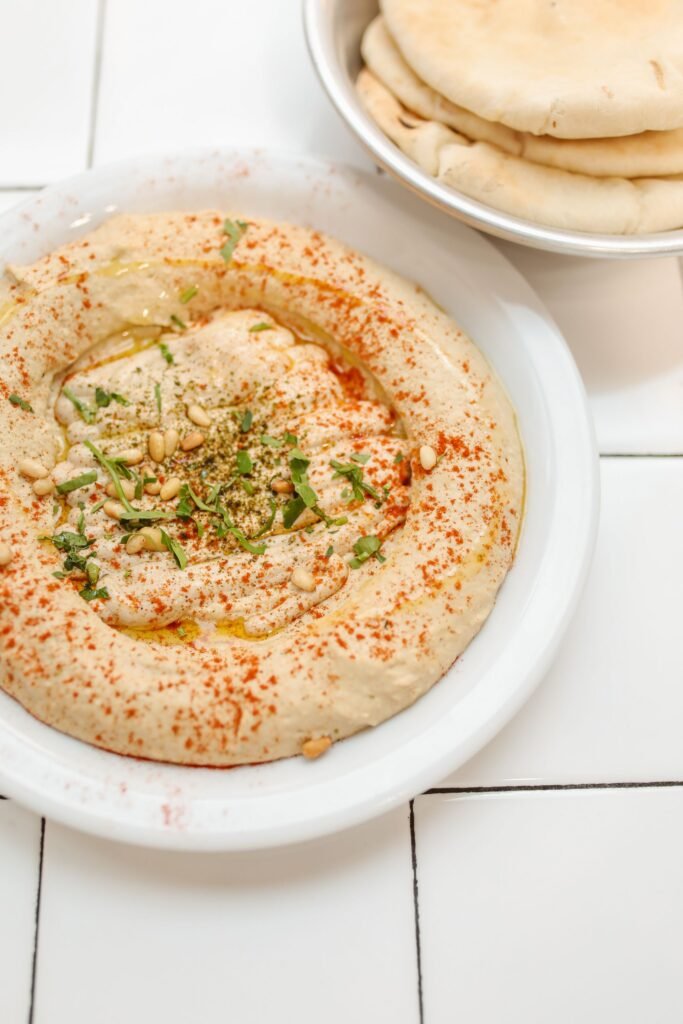 Creamy homemade hummus topped with olive oil, paprika, fresh herbs, and pine nuts, served with pita bread.